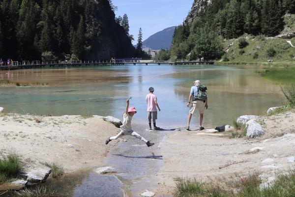 A family enjoys a sunny day by a clear mountain lake, with a child playfully jumping over a small stream